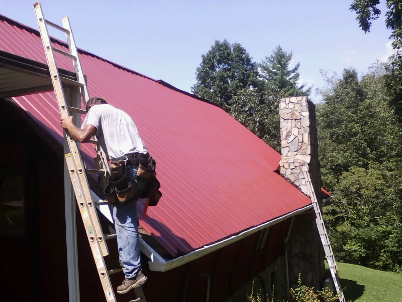 Roofer on a ladder beside a red metal roof and stone chimney — Soffit & Fascia Repair by Roofing Brothers in Morning View